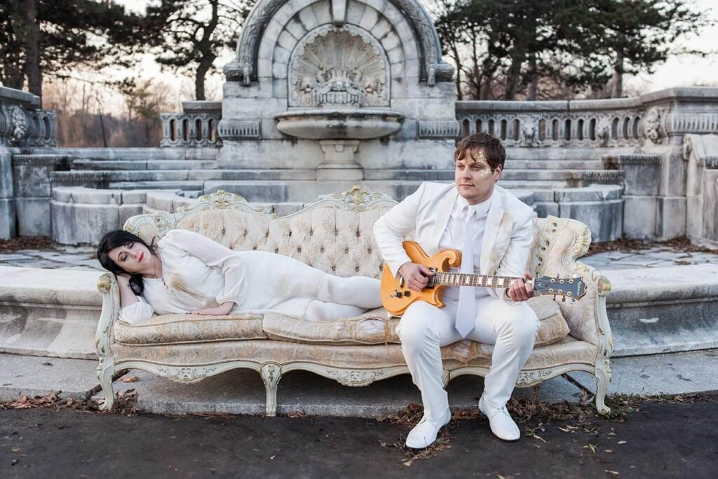 woman lying down on white vintage sofa, man sitting on sofa wearing white suit and holding a guitar, set in front of an old fountain